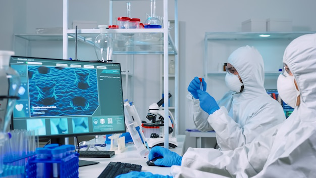 A chemist doctor in PPE working on a PC while a lab technician uses a microscope.